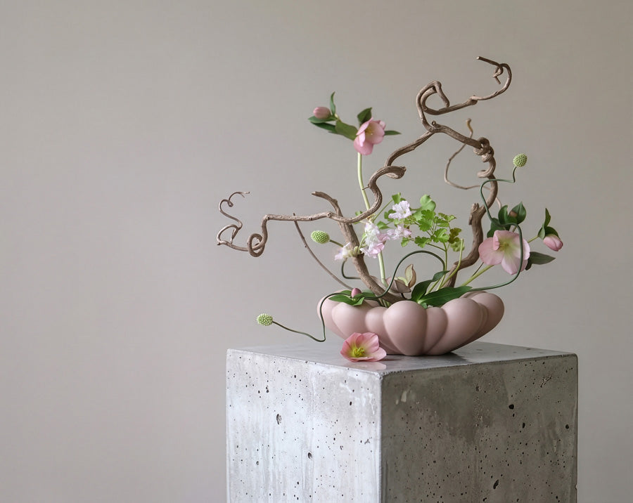 A pink Bloom Tray from 101 Copenhagen, holding an artistic floral arrangement with pink blossoms, green leaves, and twisted brown branches, displayed on a square concrete pedestal against a plain light gray wall.