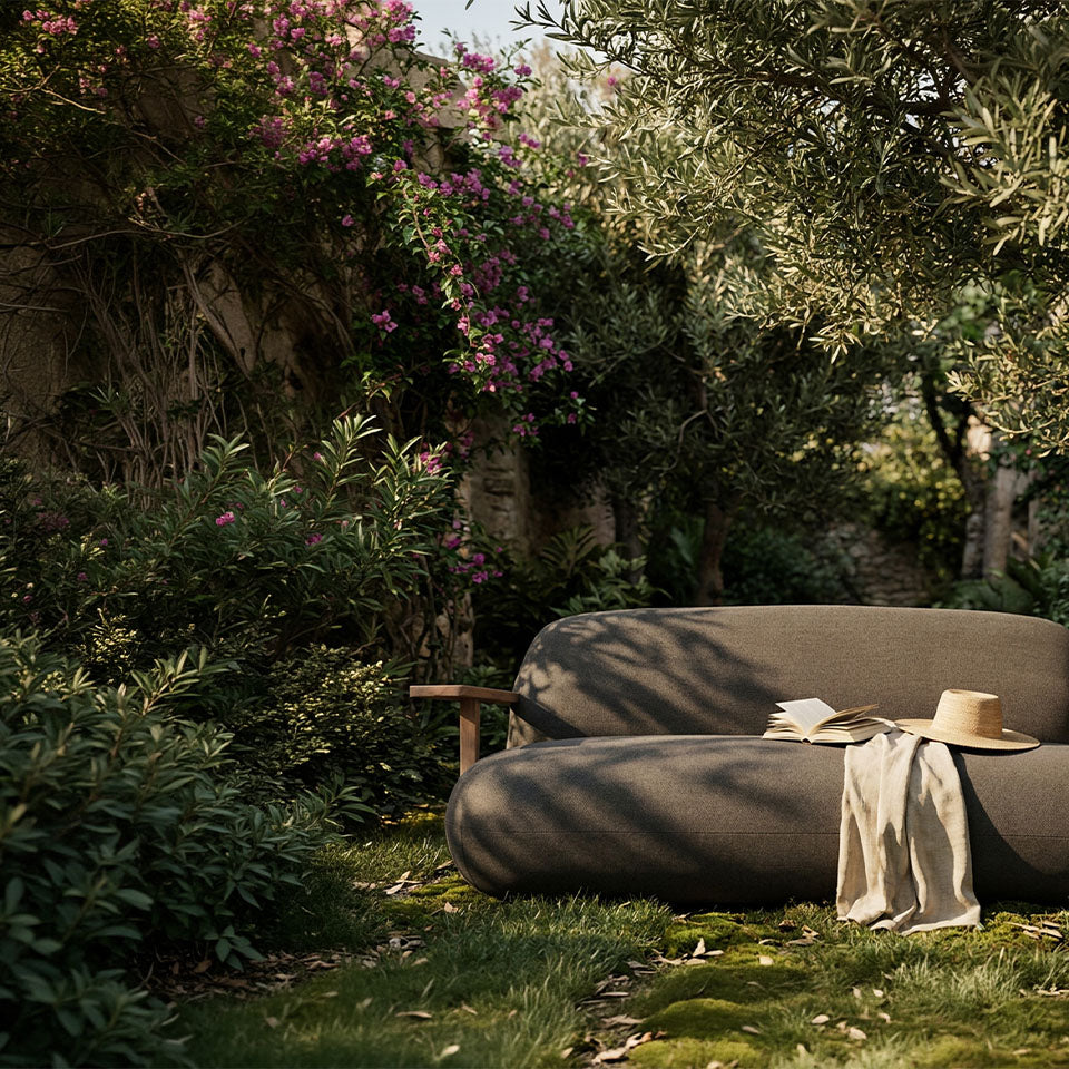 A modern outdoor lounge Toe Sofa in a lush garden setting, styled with a straw hat, a book, and a beige throw blanket.