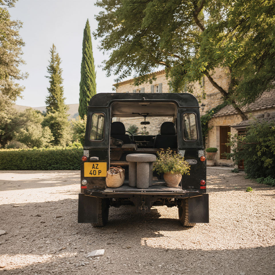 A black vintage Land Rover truck parked outdoors on a gravel driveway in front of a stone farmhouse, with its rear doors open and a Big foot stool and potted plants on the back deck.