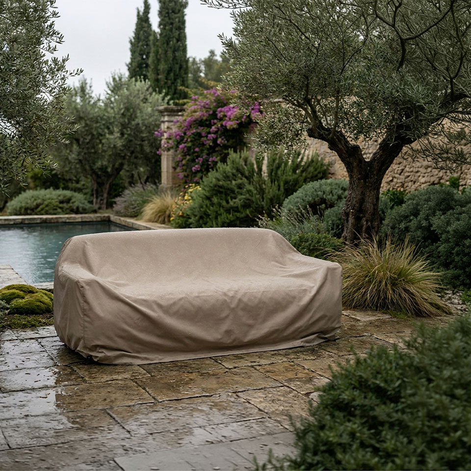 A beige patio cover sofa is shown outdoors on a stone patio next to a pool, surrounded by olive trees, shrubs, and a stone wall.