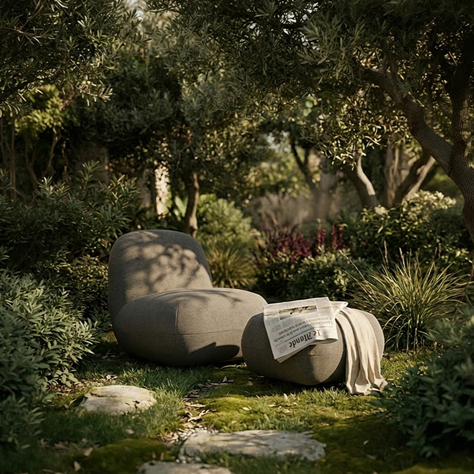 Modern organic-shaped ottoman and Toe chairs in textured taupe fabric, set in a lush, sun-dappled garden with mossy grass and olive trees. One chair is styled with a folded newspaper, and a light beige throw blanket is draped over the footrest of the other chair.