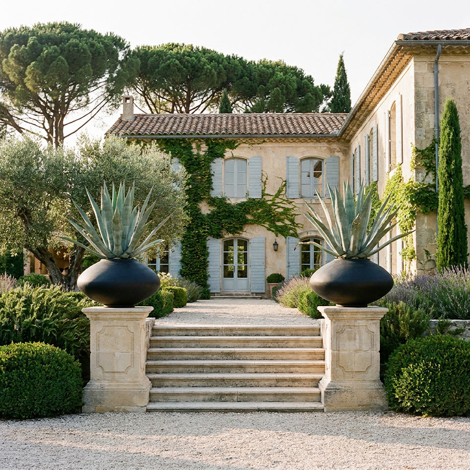 A grand stone villa with light blue shutters and ivy-covered walls, framed by tall trees and manicured shrubs. A central stone staircase is flanked by two large black Sunao Plant pots holding tall agave plants, set on a gravel foreground.