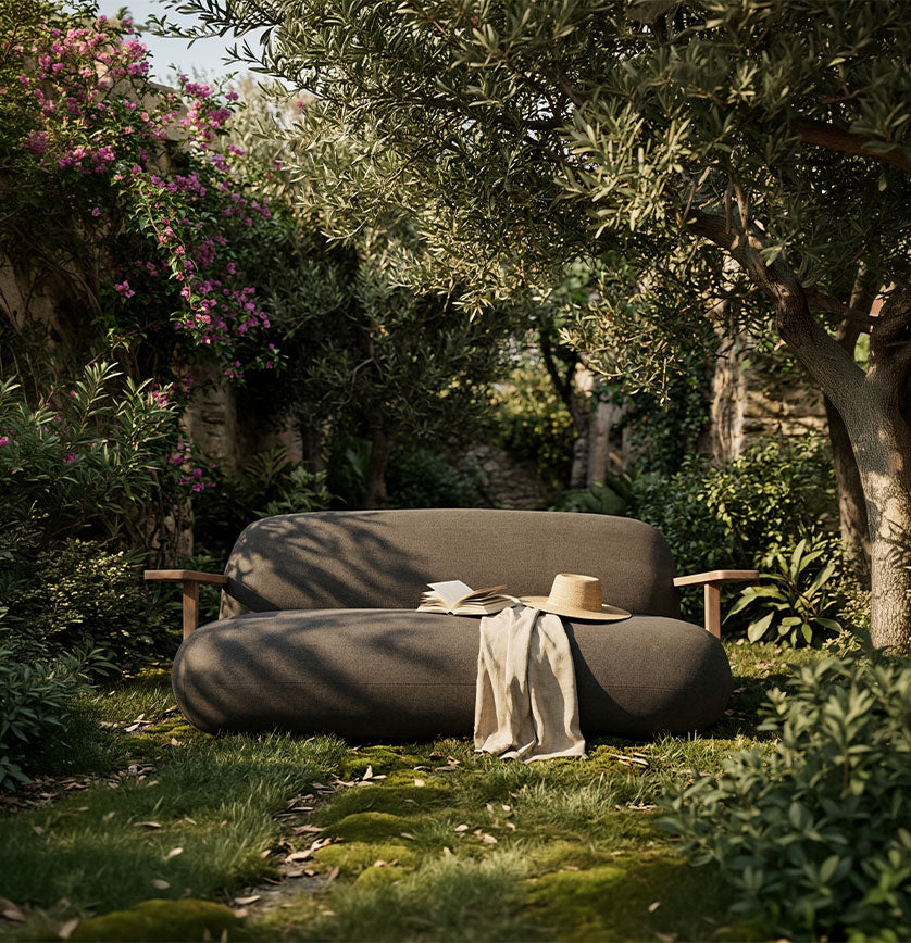 A modern curved Toe sofa in a lush garden setting, styled with a straw hat and a beige throw blanket.