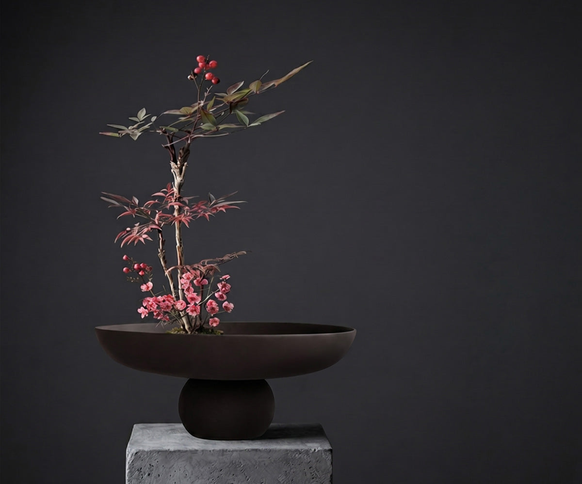 A Chocolate brown ceramic Baburu bowl, styled with a delicate bonsai tree branch featuring pink blossoms and red berries, set against a dark gray background on a concrete block.