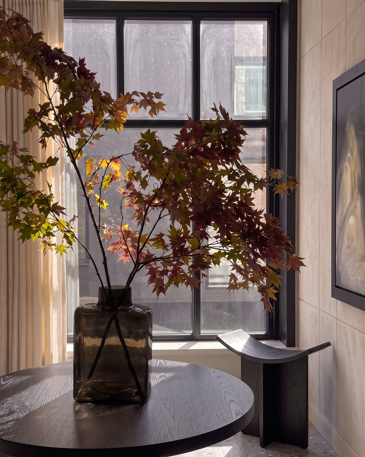 A Shin glass vase in a dark smoked forest colour, filled with autumn-colored maple leaves, displayed on a round dark wood table in a modern interior setting with a large window and a sculptural black bench.