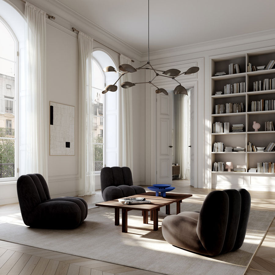 A modern living room with four dark upholstered Toe lounge chairs arranged around a wooden Plinth coffee table with Marrakech Blue Duck plates on a large cream rug. The room has high arched windows with white drapes, built-in bookshelves, light wood herringbone flooring, and a sculptural multi-arm Stingray Chandelier hanging from the ceiling.