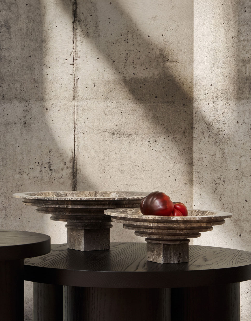 Two round Sori Trays in Silver travertine with tiered rims sit on a dark wooden table against a textured concrete wall, with one bowl holding two red tomatoes. 